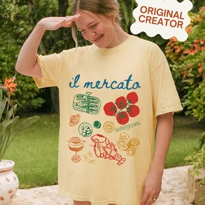 Op de afbeelding: Een geel T-shirt met een grafische print van verschillende Italiaanse voedingsmiddelen, waaronder tomaten, een kreeft en een blikje sardines. De tekst "il mercato" is in blauw op het shirt gedrukt.