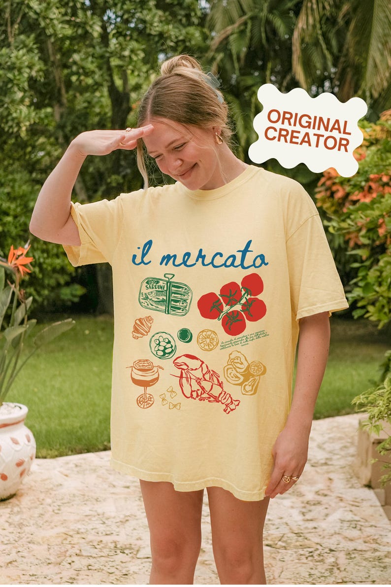 Op de afbeelding: Een geel T-shirt met een grafische print van verschillende Italiaanse voedingsmiddelen, waaronder tomaten, een kreeft en een blikje sardines. De tekst "il mercato" is in blauw op het shirt gedrukt.