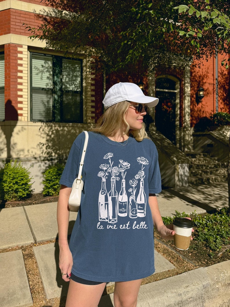 Peut inclure: Une femme portant un t-shirt bleu avec un motif blanc de bouteilles de vin et de fleurs. Le t-shirt dit "la vie est belle". Elle porte une casquette de baseball blanche et des lunettes de soleil. Elle tient une tasse de caf&eacute;.