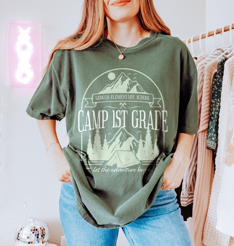May include: Green t-shirt featuring a graphic of a mountain range, a tent, and trees. The text "Gower Elementary School Camp 1st Grade" is printed on the shirt. The text "Let the adventure begin" is printed below the graphic.