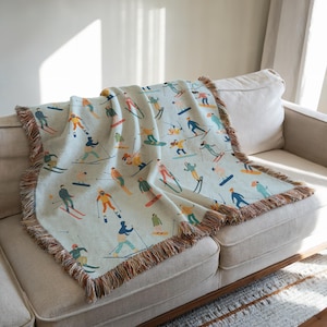 May include: A light-colored woven throw blanket featuring a repeating pattern of skiers and snowboarders in various colorful outfits. The blanket has a fringed edge in shades of brown and orange. The blanket is draped over a beige sofa.