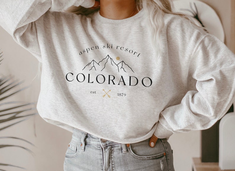K&ouml;nnte beinhalten: Ein hellgraues Sweatshirt mit dem Text "Aspen Ski Resort Colorado" in Schwarz, mit einer Berg-Grafik und dem Jahr "1879". Das Sweatshirt hat einen Rundhalsausschnitt.