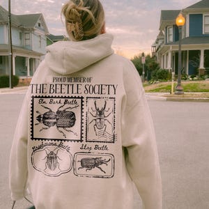 K&ouml;nnte beinhalten: Ein beiger Hoodie mit dem Text "Proud Member of The Beetle Society" und Bildern von verschiedenen K&auml;ferarten, darunter ein Borkenk&auml;fer, ein Hirschk&auml;fer und ein Nashornk&auml;fer.