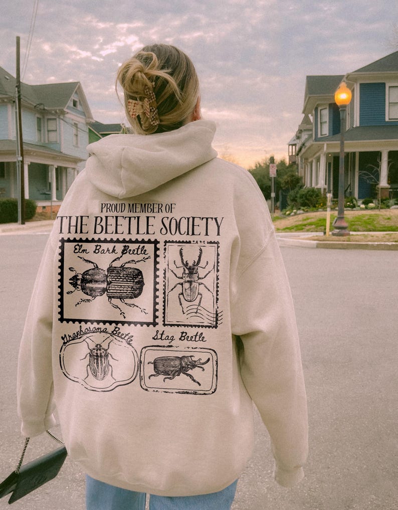 K&ouml;nnte beinhalten: Ein beiger Hoodie mit dem Text "Proud Member of The Beetle Society" und Bildern von verschiedenen K&auml;ferarten, darunter ein Borkenk&auml;fer, ein Hirschk&auml;fer und ein Nashornk&auml;fer.