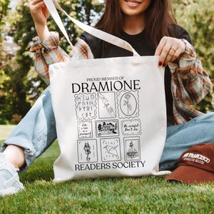 May include: A white tote bag with black text and illustrations. The text reads "Proud Member of Dramione Readers Society". The illustrations include a rose, a house, a book, and a bird.