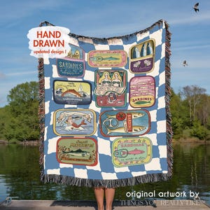 May include: A blue and white checkered throw blanket featuring colorful sardine tin illustrations. The blanket has a fringed edge and text that reads "HAND DRAWN updated design!" and "original artwork by THINGS YOU REALLY LIKE!"