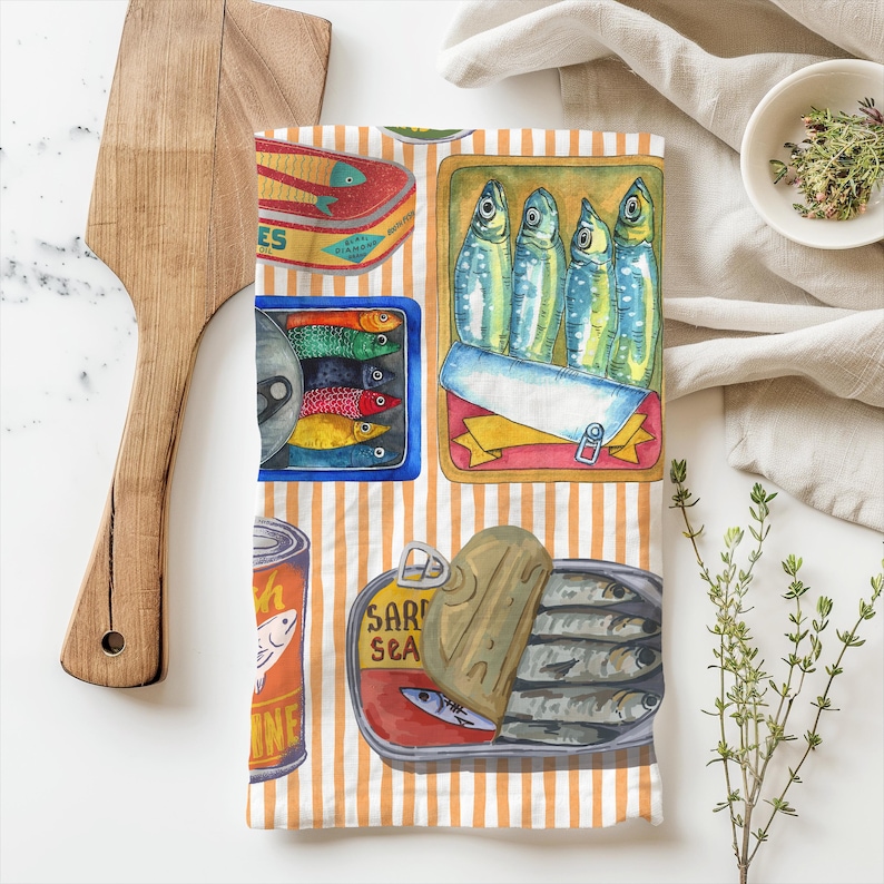 May include: A kitchen tea towel with a colourful pattern of striped orange and white background with various illustrations of canned fish. The text "Sardine Sea" is visible on one of the cans.