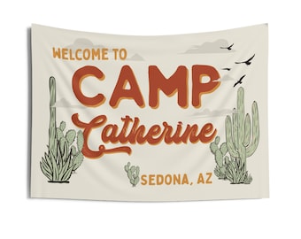 May include: A beige flag with the text "Welcome to Camp Catherine Sedona, AZ" in brown and orange lettering. The flag features a stylized illustration of cacti and birds.