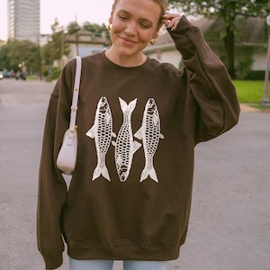 May include: A brown sweatshirt with a white graphic of three fish. The sweatshirt has long sleeves and a crew neck.