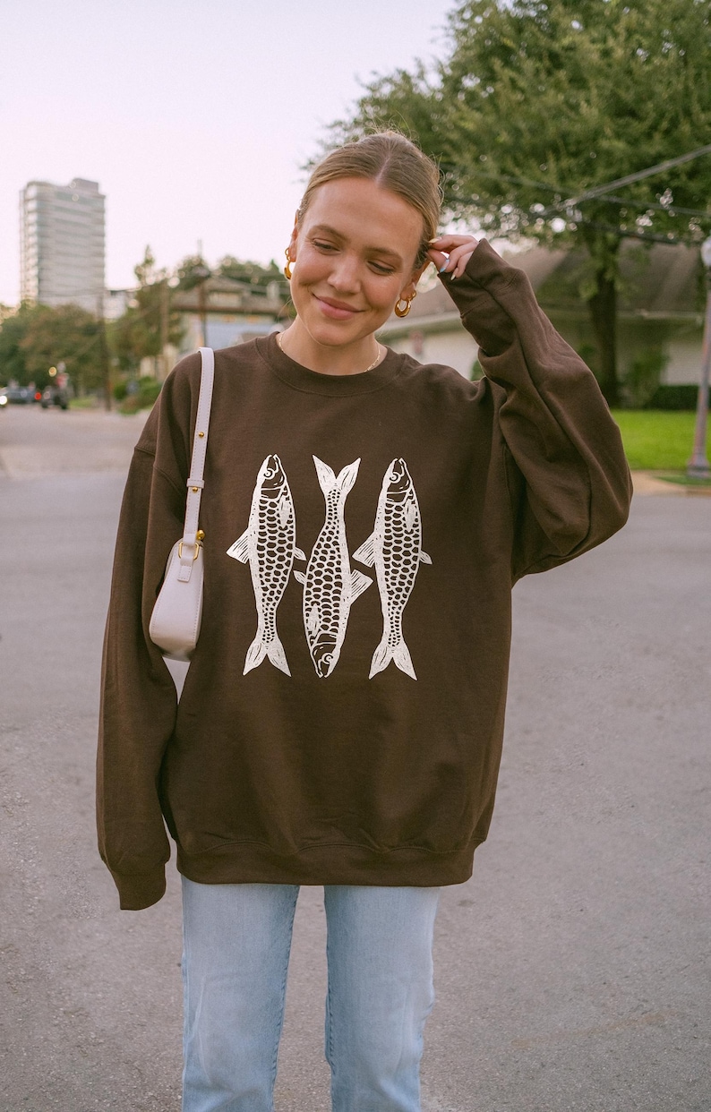 May include: A brown sweatshirt with a white graphic of three fish. The sweatshirt has long sleeves and a crew neck.