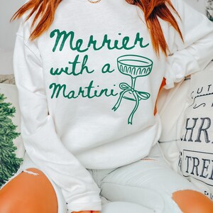 May include: White sweatshirt with green text that reads "Merrier with a Martini" and a green illustration of a martini glass with a bow.