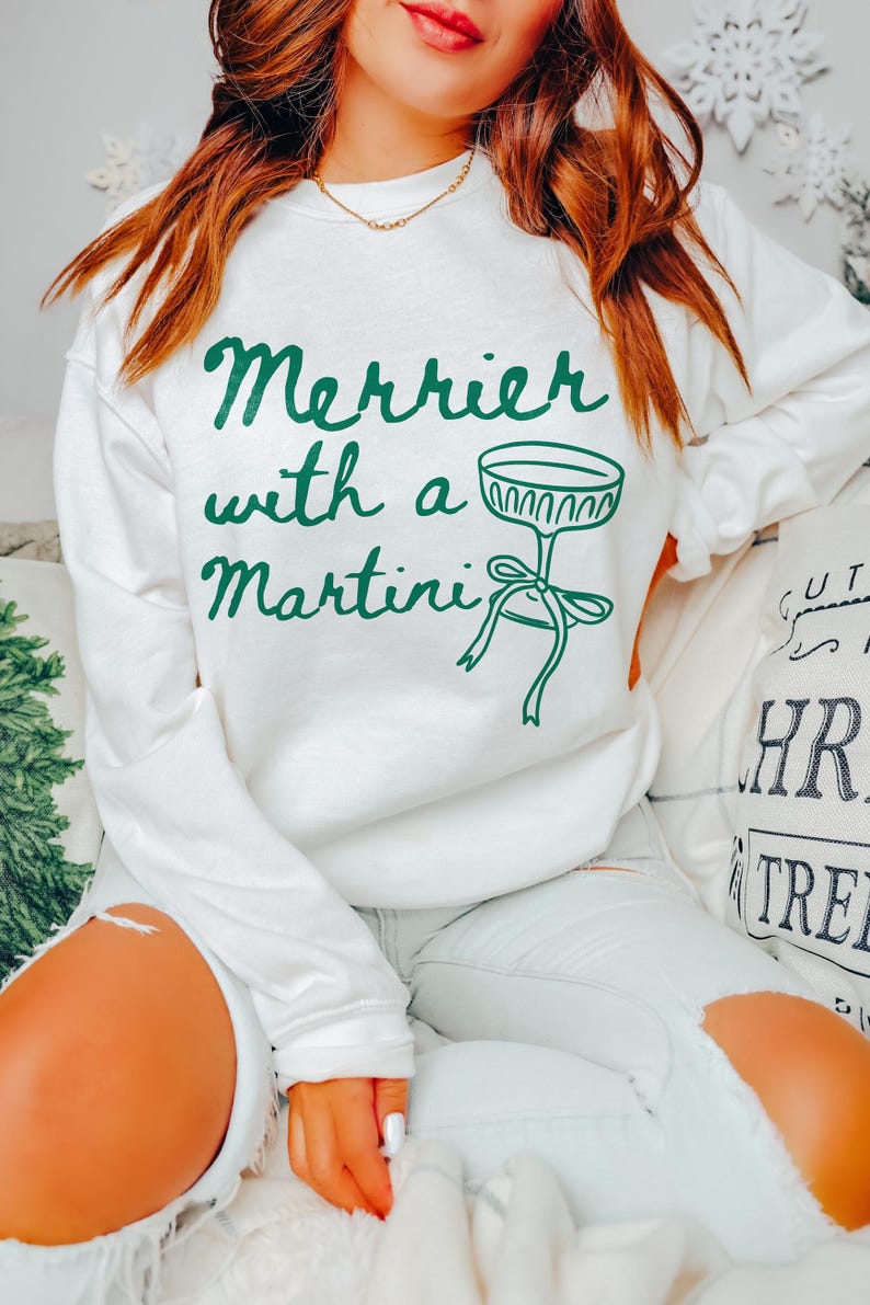 May include: White sweatshirt with green text that reads "Merrier with a Martini" and a green illustration of a martini glass with a bow.