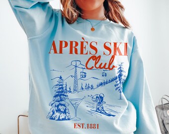 Op de afbeelding: Een lichtblauw sweatshirt met een grafische print van een skitafereel en de tekst "APRÈS SKI Club EST. 1881".