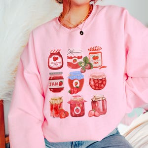 May include: Pink sweatshirt with a graphic of nine jars of strawberry jam. Each jar has a different design and is filled with red strawberries. The text "Strawberry Jam" is printed on one of the jars.