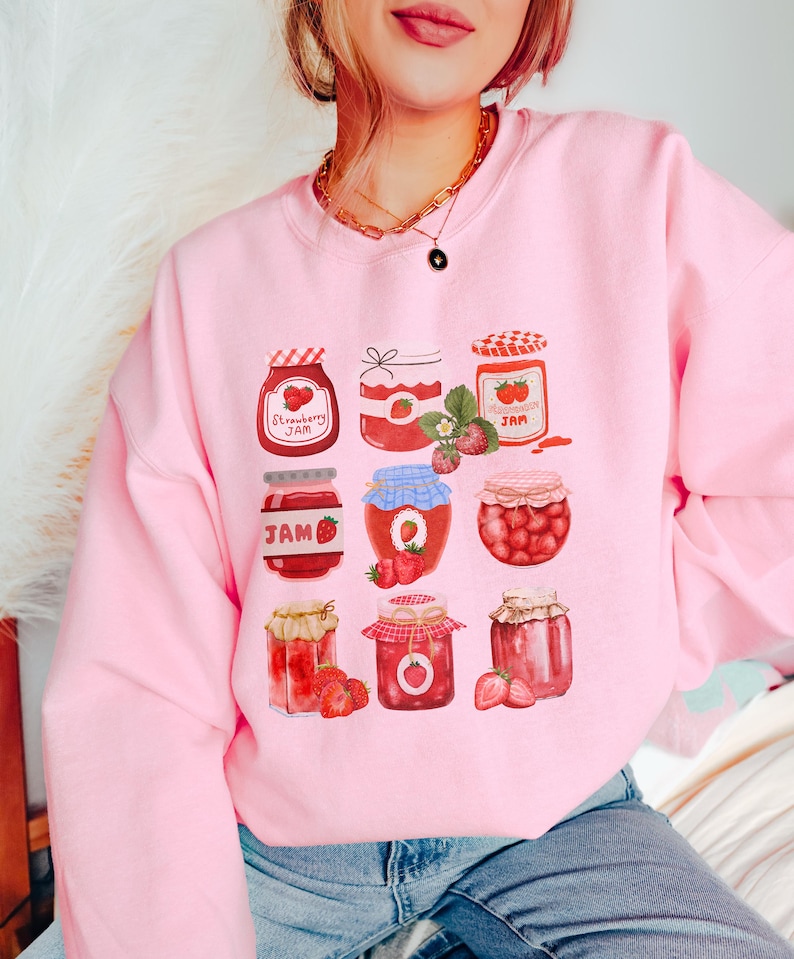 May include: Pink sweatshirt with a graphic of nine jars of strawberry jam. Each jar has a different design and is filled with red strawberries. The text "Strawberry Jam" is printed on one of the jars.