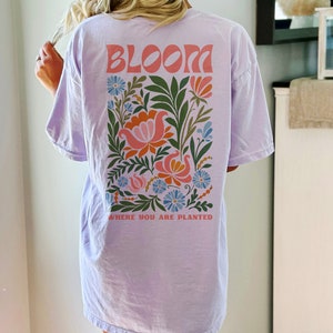 Puede incluir: Camiseta lila claro con la palabra "BLOOM" en letras coral encima de un dise&ntilde;o floral. El dise&ntilde;o incluye flores y hojas naranjas, rosas, azules y verdes. El texto "WHERE YOU ARE PLANTED" est&aacute; debajo.