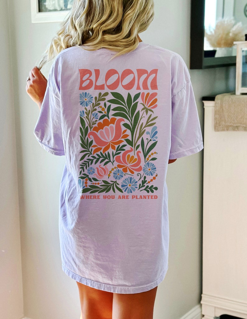 Puede incluir: Camiseta lila claro con la palabra "BLOOM" en letras coral encima de un dise&ntilde;o floral. El dise&ntilde;o incluye flores y hojas naranjas, rosas, azules y verdes. El texto "WHERE YOU ARE PLANTED" est&aacute; debajo.