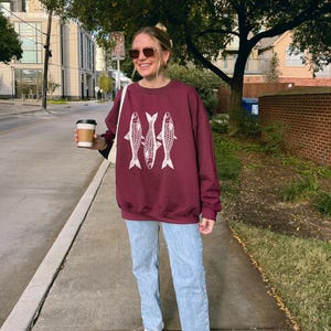 May include: A woman wearing a maroon sweatshirt with a white graphic of three fish. She is also wearing light blue jeans and white sneakers. She is holding a to-go coffee cup.