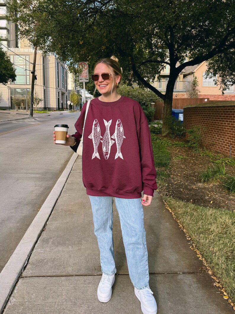 May include: A woman wearing a maroon sweatshirt with a white graphic of three fish. She is also wearing light blue jeans and white sneakers. She is holding a to-go coffee cup.