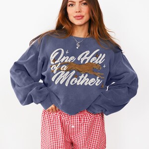 Peut inclure: Un sweat-shirt bleu avec un imprim&eacute; graphique d'un gu&eacute;pard courant et le texte "One Hell of a Mother". Le mannequin porte un short de boxe en vichy rouge et blanc.