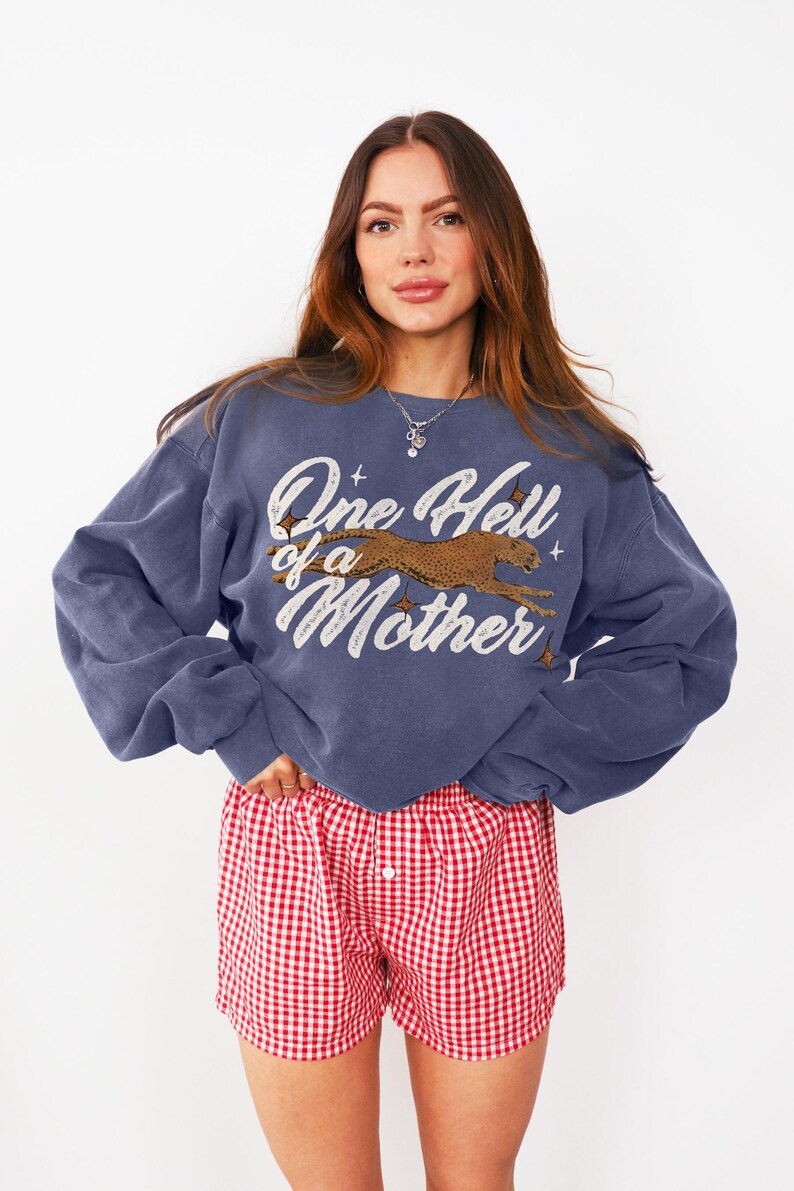 Peut inclure: Un sweat-shirt bleu avec un imprim&eacute; graphique d'un gu&eacute;pard courant et le texte "One Hell of a Mother". Le mannequin porte un short de boxe en vichy rouge et blanc.