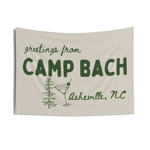 Op de afbeelding: Een beige vlag met groene tekst die "greetings from CAMP BACH Asheville, NC" luidt. Op de vlag staan ook een groene dennenboom en een groen martini-glas met een olijf.
