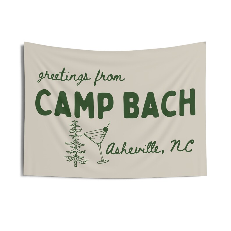 Op de afbeelding: Een beige vlag met groene tekst die "greetings from CAMP BACH Asheville, NC" luidt. Op de vlag staan ook een groene dennenboom en een groen martini-glas met een olijf.
