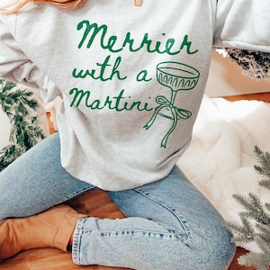 May include: A gray sweatshirt with the text "Merrier with a Martini" in green lettering and a green illustration of a martini glass with a bow.