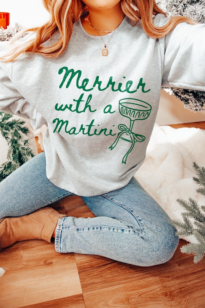 May include: A gray sweatshirt with the text "Merrier with a Martini" in green lettering and a green illustration of a martini glass with a bow.