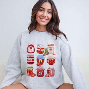 May include: A gray sweatshirt with a graphic print of nine jars of strawberry jam. The jars are decorated with red strawberries and have white lids. The text "Strawberry Jam" is printed on one of the jars.