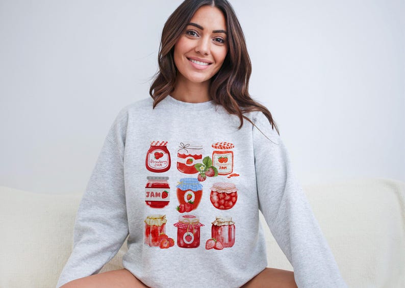 May include: A gray sweatshirt with a graphic print of nine jars of strawberry jam. The jars are decorated with red strawberries and have white lids. The text "Strawberry Jam" is printed on one of the jars.