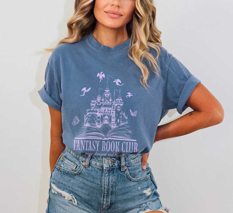 May include: A blue t-shirt with a purple graphic of a castle on an open book with dragons flying around it. The text "Fantasy Book Club" is below the graphic with the words "magic, dragons and the like" underneath.