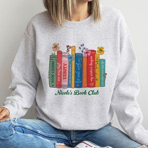 Op de afbeelding: Grijs sweatshirt met een afbeelding van een stapel kleurrijke boeken met bloemen erop. De tekst "Nicole's Book Club" is onder de boeken gedrukt.