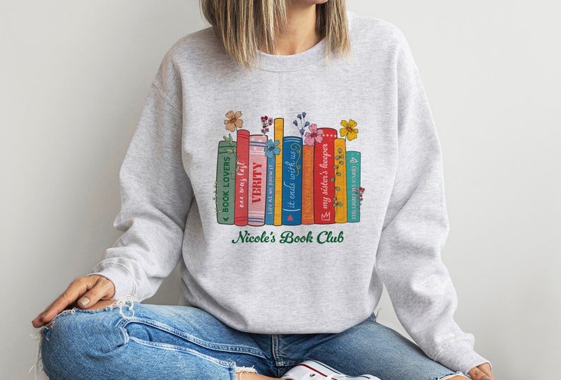 Op de afbeelding: Grijs sweatshirt met een afbeelding van een stapel kleurrijke boeken met bloemen erop. De tekst "Nicole's Book Club" is onder de boeken gedrukt.