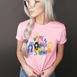 Pu&ograve; includere: Una maglietta rosa con un design grafico di donne in STEM. La grafica presenta un gruppo di donne con diversi toni della pelle e acconciature, e il testo "Women in STEM" &egrave; scritto in un font colorato.