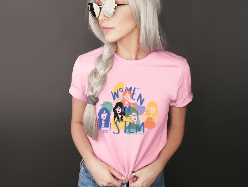 Pu&ograve; includere: Una maglietta rosa con un design grafico di donne in STEM. La grafica presenta un gruppo di donne con diversi toni della pelle e acconciature, e il testo "Women in STEM" &egrave; scritto in un font colorato.