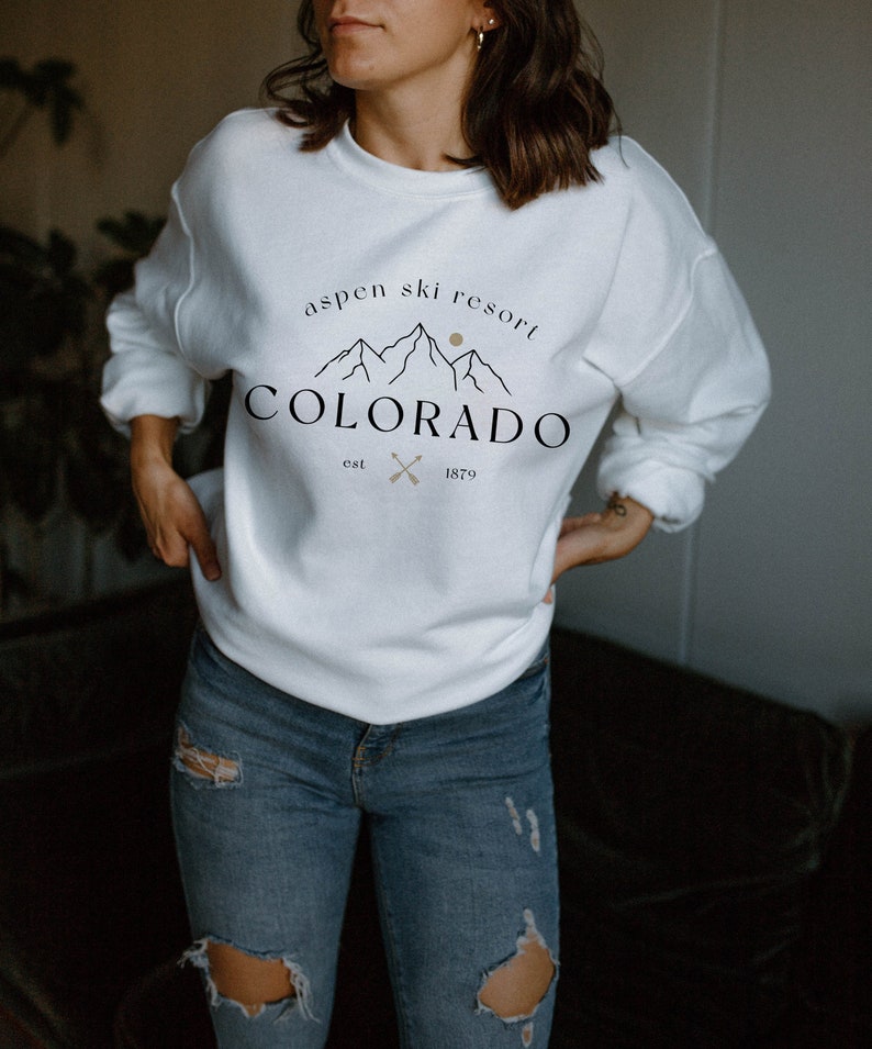 K&ouml;nnte beinhalten: Wei&szlig;es Sweatshirt mit dem Text "Aspen Ski Resort Colorado" in Schwarz und Beige. Das Design umfasst Berggrafiken, eine Sonne und gekreuzte Pfeile. Das Sweatshirt ist ein l&auml;ssiger Stil.