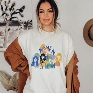 Pu&ograve; includere: Una felpa bianca con una grafica colorata di donne in STEM. La grafica include il testo "Women in STEM" e presenta illustrazioni di donne con diversi stili di capelli e tonalit&agrave; della pelle.