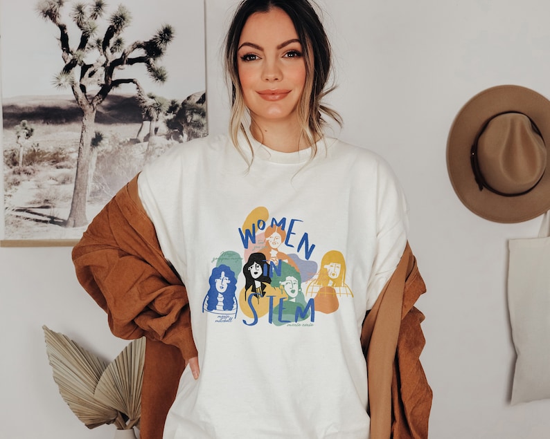 Pu&ograve; includere: Una felpa bianca con una grafica colorata di donne in STEM. La grafica include il testo "Women in STEM" e presenta illustrazioni di donne con diversi stili di capelli e tonalit&agrave; della pelle.