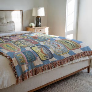 May include: A woven throw blanket featuring a repeating pattern of vintage sardine can labels in various colors. The blanket has a blue and white checkered background and a fringed edge. The blanket is draped over a bed.