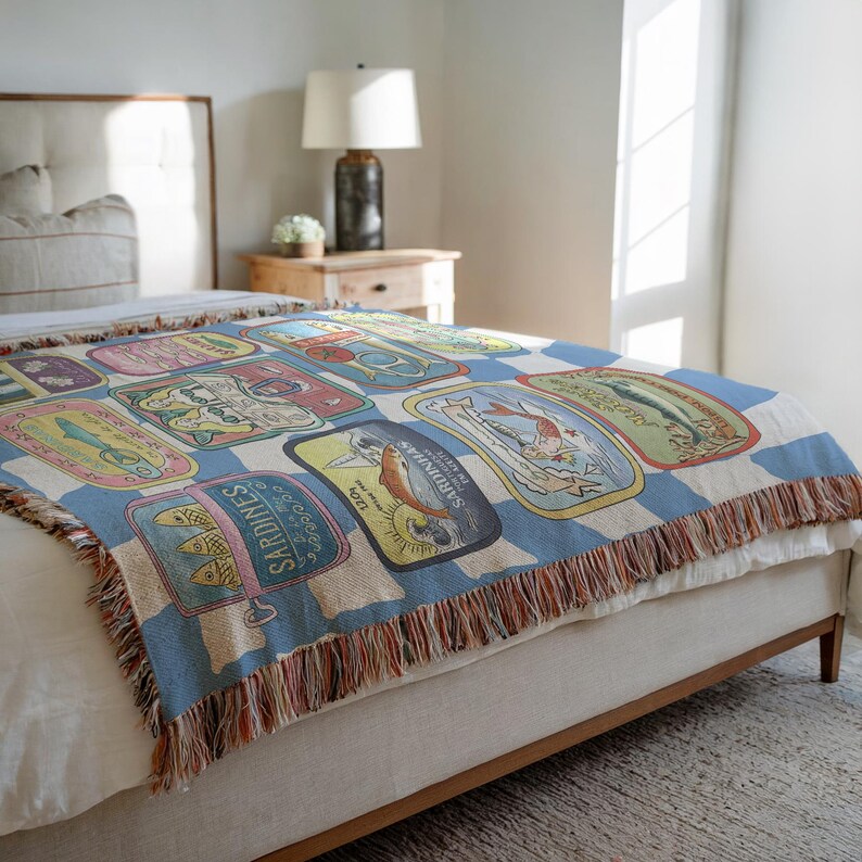 May include: A woven throw blanket featuring a repeating pattern of vintage sardine can labels in various colors. The blanket has a blue and white checkered background and a fringed edge. The blanket is draped over a bed.