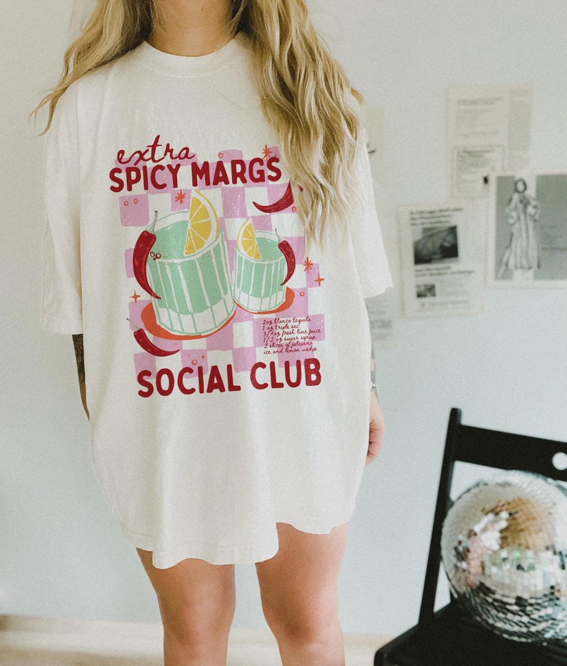 May include: White oversized t-shirt with a pink and red graphic design featuring two glasses of a spicy margarita cocktail. The text on the shirt reads "extra spicy margs social club".