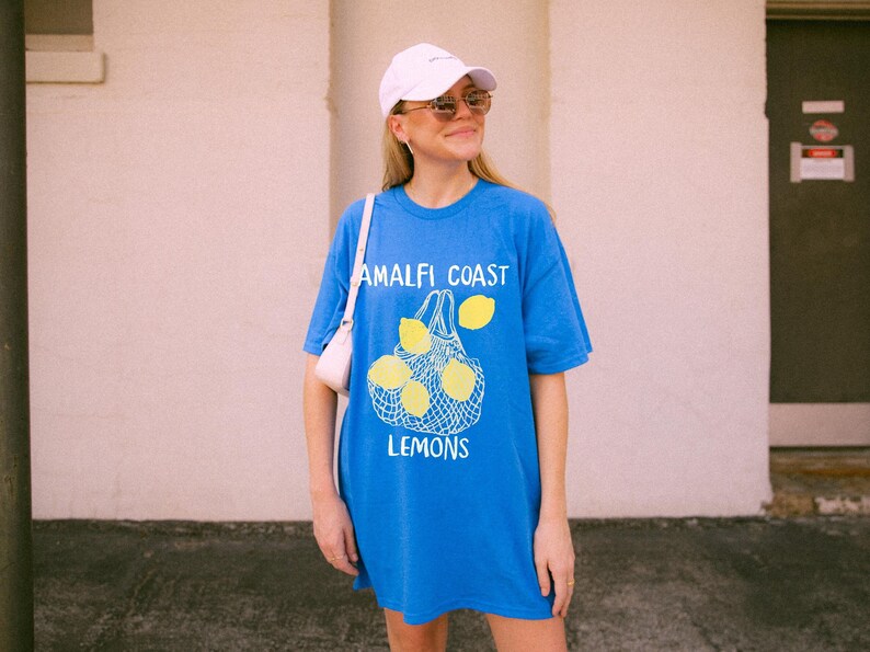 May include: A woman wearing a white baseball cap and sunglasses is standing in front of a white wall. She is wearing a blue t-shirt with a graphic of lemons in a mesh bag and the text "Amalfi Coast Lemons".