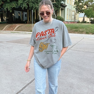May include: A grey t-shirt with a graphic of a fork and spaghetti noodles. The text on the shirt reads "Pasta Lover Club" and "consumed daily 7pm - 10pm".
