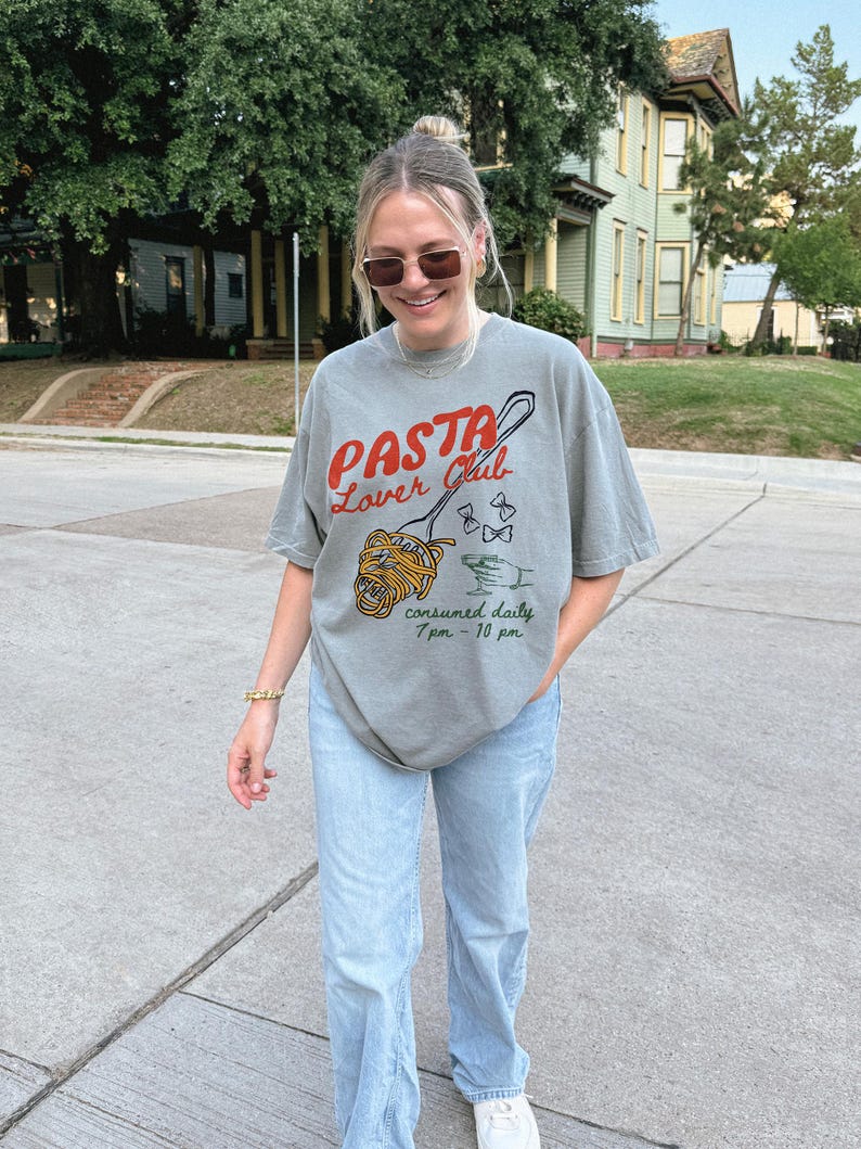 May include: A grey t-shirt with a graphic of a fork and spaghetti noodles. The text on the shirt reads "Pasta Lover Club" and "consumed daily 7pm - 10pm".
