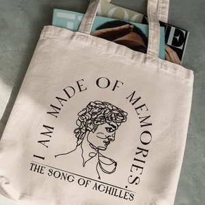 May include: A beige tote bag with black text that reads "I am made of memories. The song of Achilles.". A line drawing of a person's head is in the center of the text.