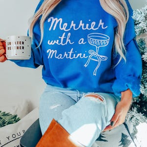 May include: A woman wearing a blue sweatshirt with the text "Merrier with a Martini" and a white martini glass graphic. She is holding a white mug with gold polka dots and the text "Sweater Weather".