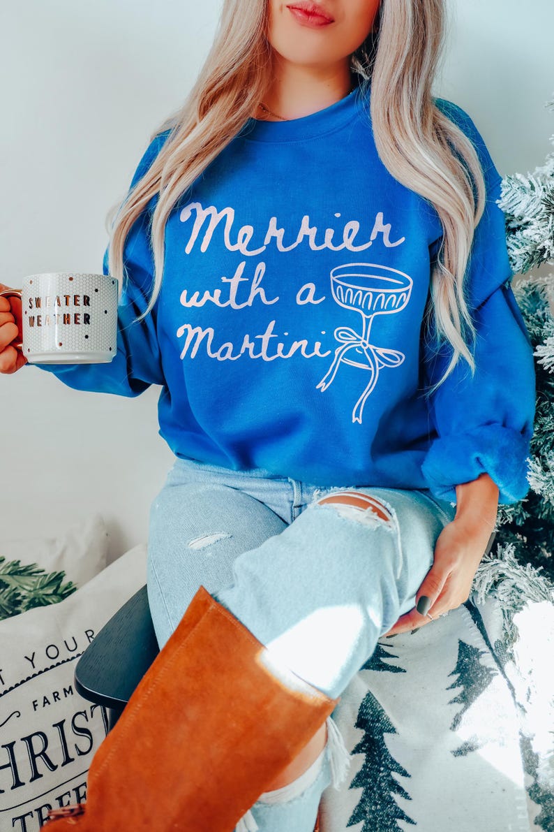 May include: A woman wearing a blue sweatshirt with the text "Merrier with a Martini" and a white martini glass graphic. She is holding a white mug with gold polka dots and the text "Sweater Weather".