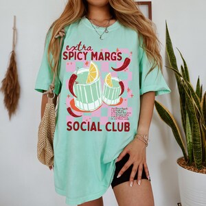 May include: A turquoise blue t-shirt with a graphic of two glasses of margaritas with limes and red chili peppers. The text "extra spicy margs social club" is printed on the shirt.