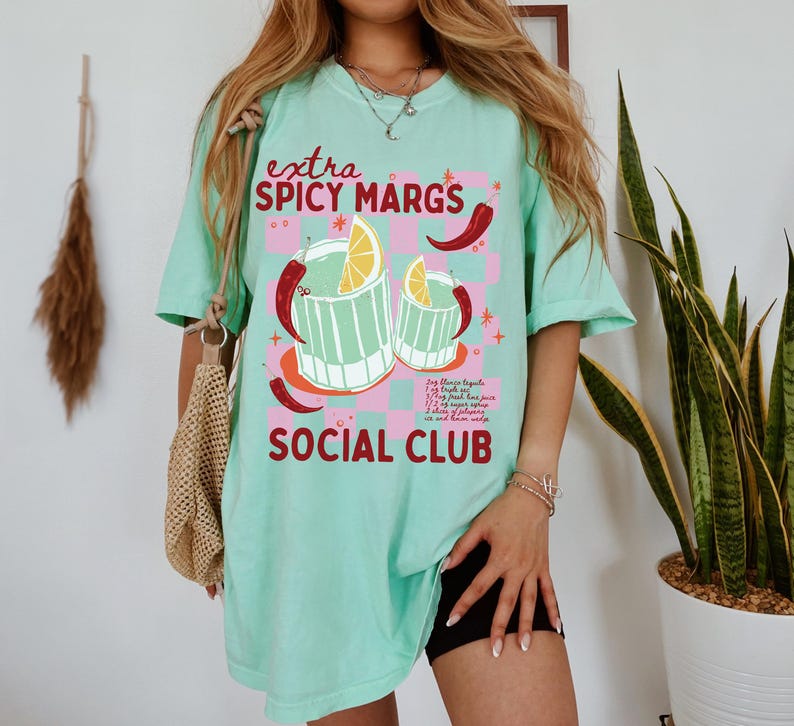 May include: A turquoise blue t-shirt with a graphic of two glasses of margaritas with limes and red chili peppers. The text "extra spicy margs social club" is printed on the shirt.